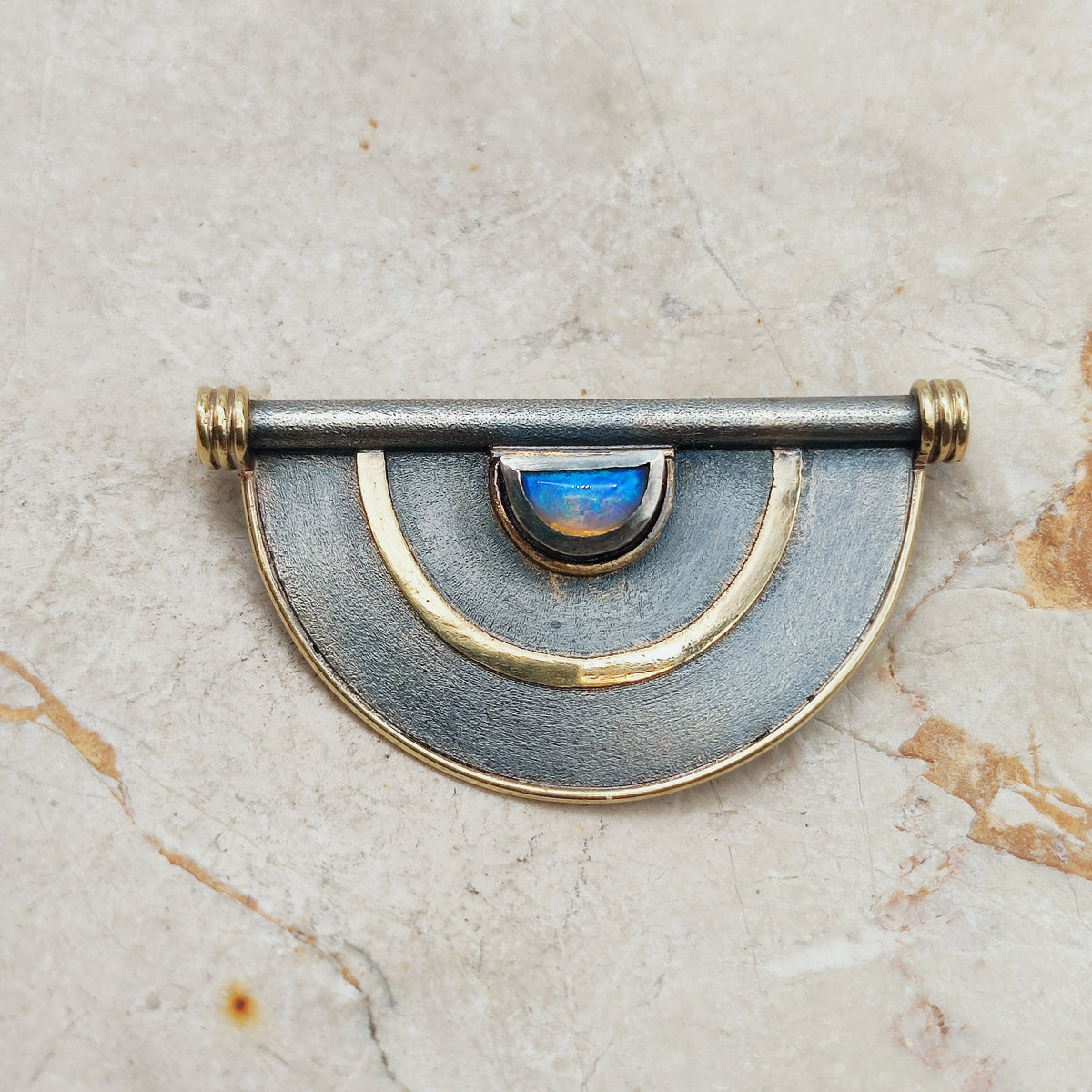 Geometrische halfronde broche in donker zilver met gouden lijnen en een blauw oplichtende opaal
